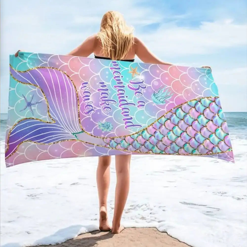 A woman standing on a beach holding an Ocean Queens colorful towel with a mermaid tail design and the text "Be a mermaid and make waves.